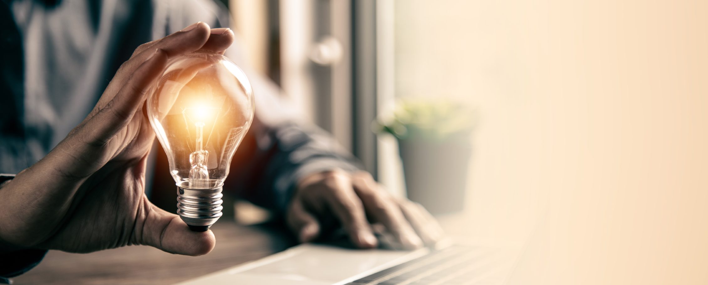 A person cradling a lit incandescent light bulb in their hands at a sunlit desk.